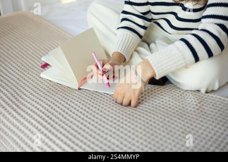 A woman wearing a striped sweater writes in a notebook using a pink pen while sitting on a bed The scene conveys relaxation, creativity, and a cozy at Stock Photo
