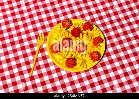 A vibrant yellow plate filled with colorful flowers rests on a classic red and white checkered tablecloth. A yellow fork complements the vivid picnic Stock Photo