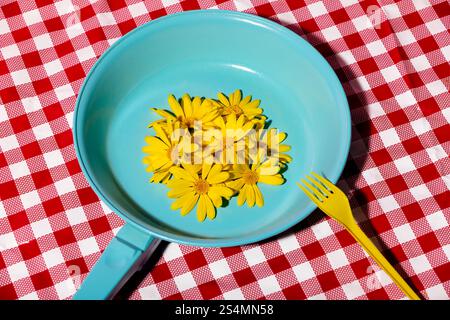 A vibrant scene featuring bright yellow flowers arranged in a blue pan, accompanied by a yellow fork, set against a red checkered tablecloth backgroun Stock Photo