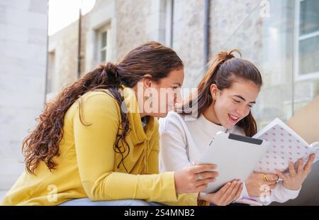 Two students enjoy a study session outside on a sunny day, using a tablet and a notebook They appear engaged and happy, back dropped by modern archite Stock Photo