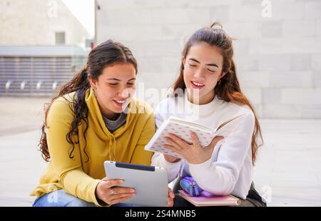 Two students sit together outdoors, enjoying a collaborative study session One holds a tablet, while the other reads a book, fostering learning and co Stock Photo