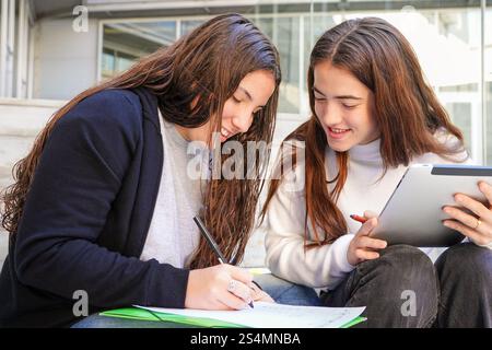 Two students are sitting outdoors, working together on a project One is writing on paper, while the other holds a tablet, indicating teamwork and digi Stock Photo