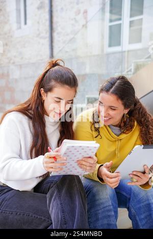 Two students sit on stairs, engaged in study and discussion One takes notes in a notebook, while the other uses a tablet Both seem focused and collabo Stock Photo