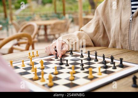 A close-up view of a chess game being played outdoors on a wooden table The chessboard is set with black and white pieces, surrounded by a relaxed, su Stock Photo