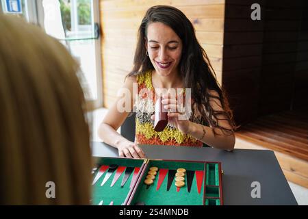 A smiling woman focuses on rolling dice while playing backgammon at a table, creating a warm and engaging atmosphere in an indoor setting The wooden b Stock Photo