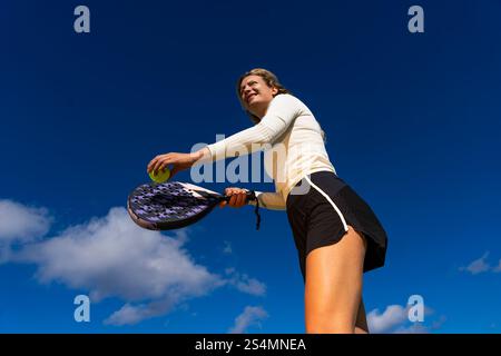 From below a young woman in sportswear is captured in the act of playing tennis against a bright blue sky background, focusing intently on her next sh Stock Photo