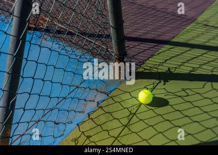 Vibrant sunlight casts intricate shadows over a tennis ball beside a net on a cracked court surface, emphasizing decay and neglect in sports facilitie Stock Photo
