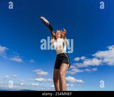 From below a young woman is captured in action while playing tennis outdoors, with a clear blue sky and distant mountains as her background. She is lo Stock Photo