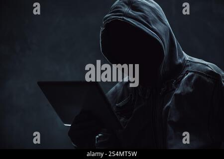 Hooded computer hacker using digital tablet, selective focus Stock Photo