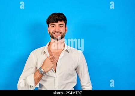 Portrait of a confident businessman smiling and opening his white shirt, showing his chest, on a vibrant blue background Stock Photo