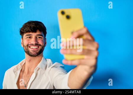 Happy businessman smiling and taking a selfie with his yellow smartphone on a vibrant blue background Stock Photo