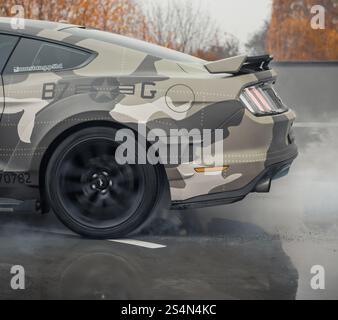 eige camo Ford Mustang doing a burnout. Rear side view of american pony car burning rubber on wet concrete surface. Stock Photo