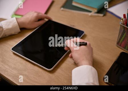 Hands holding stylus and interacting with black screen tablet, with office supplies and notebooks placed on the desk. Stock Photo