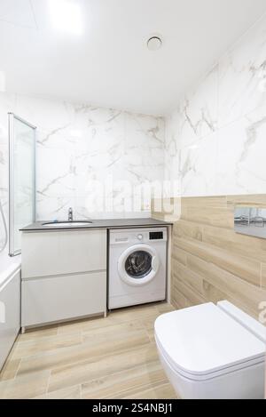 Bright and contemporary bathroom featuring a washing machine, sleek marble walls, and warm wooden floor panels Stock Photo