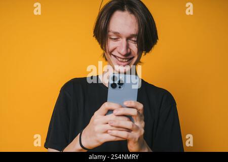 A Young Man Is Cheerfully Smiling While Using His Smartphone Against A Bright Yellow Background Stock Photo