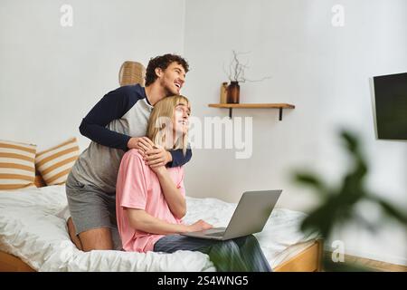 Two men share a loving embrace while using a laptop in a comfortable bedroom setting. Stock Photo
