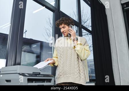 A young man in a sweater vest operates a copier while on the phone, reflecting modern work life. Stock Photo