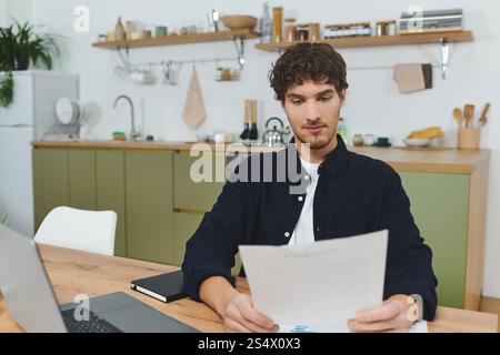 Curly-haired man sits at a wooden table in a stylish apartment, studying important papers. Stock Photo