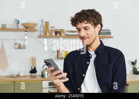Curly-haired man in a stylish shirt smiles as he scrolls through his smartphone at home. Stock Photo