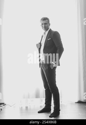 Black and white photo of handsome stylish man in suit posing against big window at modern interior Stock Photo