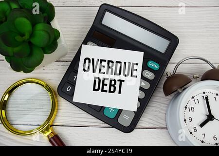 Overdue Debt text message on sticky note with magnifying glass, alarm clock and calculator on wooden background Stock Photo