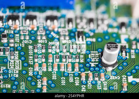 White electrolytic capacitor on printed circuit board in blurry electrotechnical background. Closeup of green blue texture with electronic components. Stock Photo