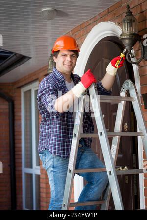 Male electrician in hardhat standing stepladder and repairing lamp on house Stock Photo