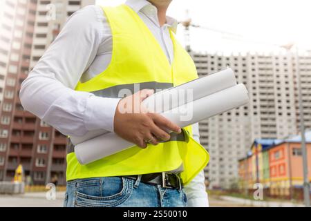 Closeup image of engineer in safety vest holding rolled blueprints Stock Photo