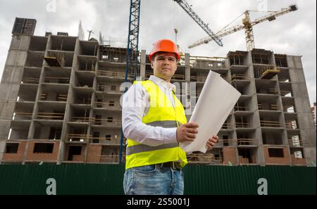 Young smiling engineer on building site checking plans and blueprints Stock Photo