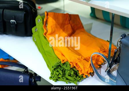 Lush green and bright orange fabrics are folded and showcased on a white shelf in a lively market. The atmosphere buzzes with energy as shoppers explo Stock Photo