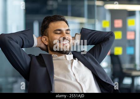 A well done man rests with his hands behind his head, dreams and thinks about the future results of his achievement. A businessman at his workplace inside the office works closeup in a business suit Stock Photo