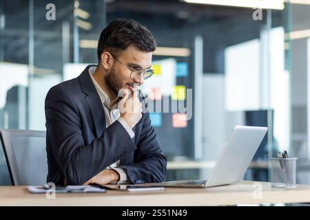Thinking businessman at workplace inside office, man in business suit working with laptop, reading online report, and preparing presentation investment project. Stock Photo