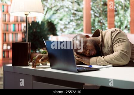 Small business owner feels tired and takes a nap while doing remote work, sleeping on his home desk. Stressed overworked freelancer falling asleep at the office setup, financial operations. Stock Photo