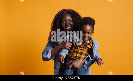 Joyful african american mother carrying her adorable child on camera, holding him close and smiling in the studio. Happy mommy and cute little boy bonding together, loving parent. Camera B. Stock Photo