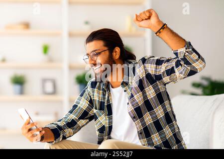Emotional long-haired indian guy hipster sitting on couch, using smartphone Stock Photo