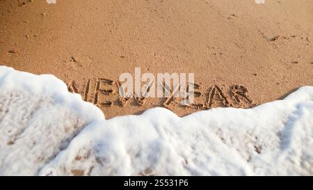 Closeup image of sea waves rolling over words New Year written on wet sand on the beach. Concept of winter holidays, Christmas and tourism. Closeup Stock Photo