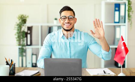 Young Arab guy teacher with laptop teaching Turkish online from home office, waving at webcam, greeting students Stock Photo