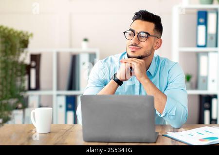 Dreamy Arab guy in glasses sitting at laptop at office, looking aside, taking break from online job Stock Photo