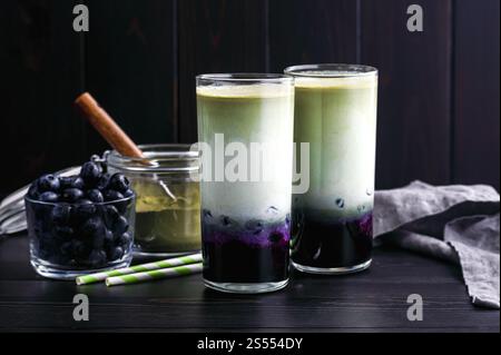 Iced Blueberry Matcha Lattes in Tall Glasses: Cold lattes made of layered green tea, milk, and blueberries with paper straws and ingredients nearby Stock Photo