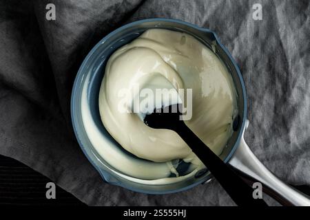 Melted White Chocolate in a Small Saucepan: Melted white chocolate mixed with a silicone spatula in a small non-stick pot overhead close-up Stock Photo