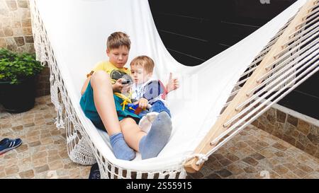 Toned image of teenage boy lying with his little brother in hammock and using smartphone Stock Photo