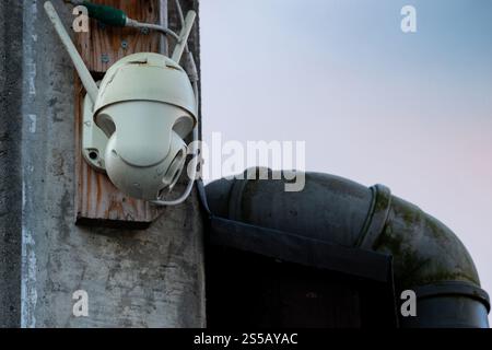 A unique surveillance camera is mounted on a wooden board attached to a concrete wall. In the background, a large pipe is visible against the sky, creating an industrial atmosphere. Stock Photo
