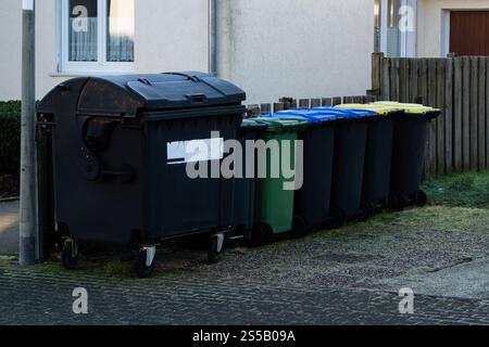 Different colored waste bins are neatly arranged along the side of a residential street. These containers include black, green, blue, and yellow bins for recycling and waste disposal. Stock Photo