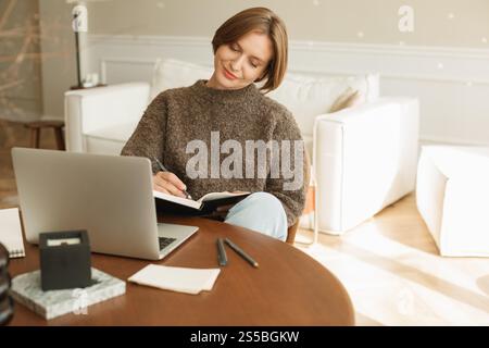 Young woman sitting at her dining table using her laptop and writing in a note pad Stock Photo
