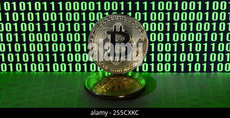 A pair of bitcoins lies on a cardboard surface on the background of a monitor depicting a binary code of bright green zeros and one units on a black Stock Photo