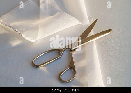 A pair of scissors is on a white cloth. The scissors are silver and have a sharp point Stock Photo