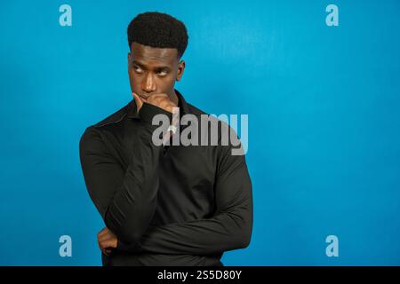 Young man in black long-sleeved shirt is holding his chin, lost in thought, against a vibrant blue backdrop Stock Photo