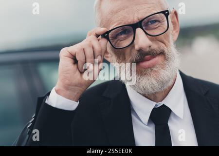 Mature businessman in formal suit adjusting glasses near car on city street Stock Photo