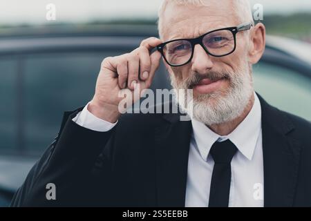 Confident senior businessman in formal suit adjusting glasses near car on city street Stock Photo
