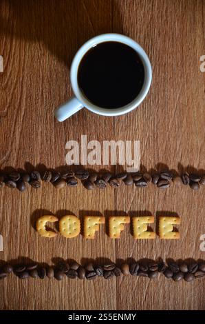 Still life with a small white cup of hot coffee and a salt cracker in the form of letters forming the word Coffee", framed by rows of coffee beans on Stock Photo
